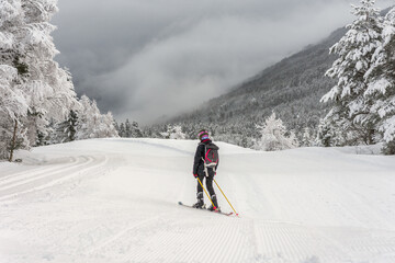 Woman with black ski wear, helmet and backpack skiing down the top of the mountain in ski centre, stryn with clouds and trees in the background, rear view, horizontal