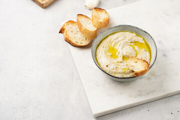 Traditional dish of arabic cuisine baba ganoush made of roasted eggplant, tahin, cilantro, garlic, olive oil, lemon juice with toasted baguette croutons on grey background, white marble board