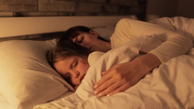 Young mother and little daughter sleeping in bed together. Mom hugging her daughter and having sweet dreams in cozy bedroom. Happy family. Sleeping together