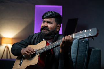 Obraz premium Young beardman playing guitar while sitting on sofa at home - concept of musician, artist and hobbies.