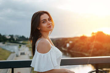 Portrait of pretty young brunette woman standing on bridge against backdrop of sunset. Side view of smiling model looking away outdoors. Beauty and fashion, lifestyle concept