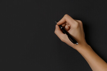 Woman hand holding pen and writing on black background, top view.