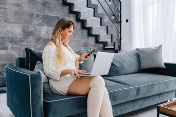 Blond attractive woman sitting blue sofa pillow remote distance work laptop, talking smartphone. Stairs, wooden table with plant pot, plate interior. Wearing white wool half hose sweater dress