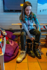 Woman with brown hair preparing for skiing, looking down while trying on one black ski shoes inside a building, vertical