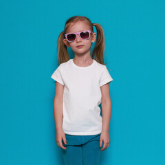 Fototapeta premium White t-shirt mockup, little european girl in summer clothes, sunglasses on a blue background