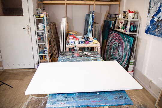 White blank canvas lying on table in arts work room with multiple paintings, tools and equipment and cans and jars with paint