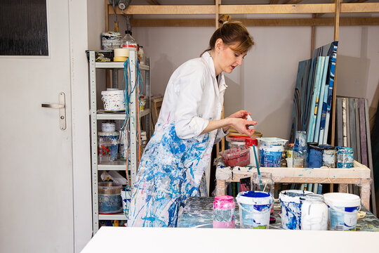 Side view of woman painter working with tools standing by table with cans and jars with paint in arts work room