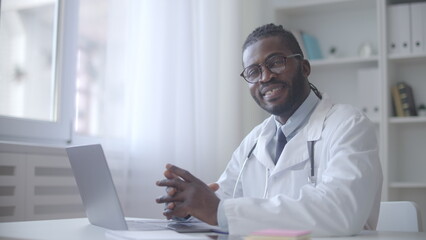 Confident general practitioner looking at camera and smiling, sitting in his office, online consultations