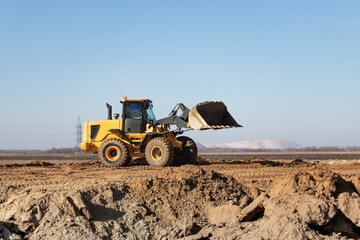 Obraz premium Bulldozer or loader moves the earth at the construction site against the blue sky. An earthmoving machine is leveling the site. Construction heavy equipment for earthworks.