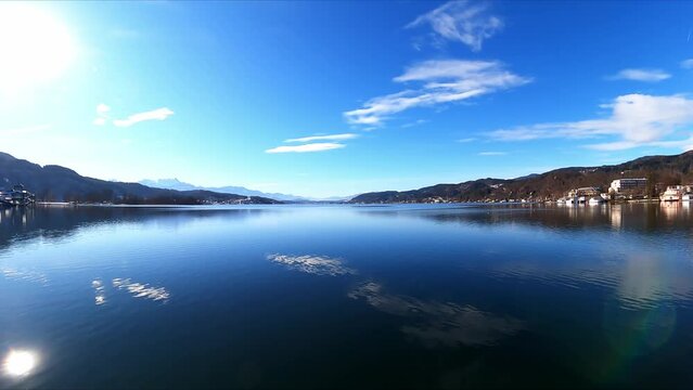 A panoramic view on the calm surface of the Woerthersee Lake in Austria. The lake is surrounded by high mountains. Small villages at the shores, and a small water park with slides. Soft clouds
