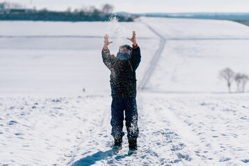 ein Kind wirft Schnee in die Luft vor einer weißen Winterlandschaft bei strahlenden Sonnenschein
