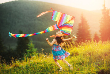 Little girl playing with colored striped rainbow kite at sunset summer day against background of beautiful mountain landscape with peaks, fir trees. Baby wearing hat and dress. Happy summer vacation