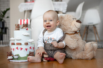 Smiling little boy in white T-shirt and checkered overall sitting on floor near two-tiered birthday cake and teddy bear.