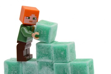 Naklejka premium LEGO Minecraft figure of Alex is building some wall from green coloured scented stearine wax cubes, white background.