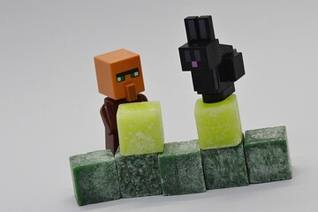 Fototapeta premium LEGO Minecraft figure of villager is placing light green scented wax cubes on wall of dark green cubes, black Minecraft rabbit is watching him from top of the light green cube.