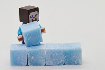 Naklejka premium LEGO Minecraft figure of Steve is building a wall from light blue cubes made of scenty stearine wax used for aromatherapy, light grey background.