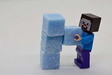 Fototapeta premium LEGO Minecraft figure of Steve is building a column from light blue cubes made of scented stearine wax, light grey background.