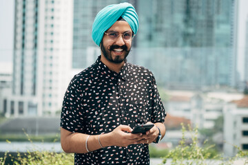 Portrait of Happy Indian Businessman Using Mobile Phone Outdoors. 
Cheerful smiling business man with turban typing text message on his phone and looking at camera while standing in the city.