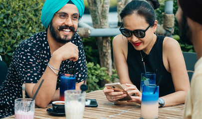 Three Multi Ethnic Friends Having Fun in an Outdoor Cafe. 
Cheerful happy friends talking and laughing together while drinking cocktails and using mobile phone.