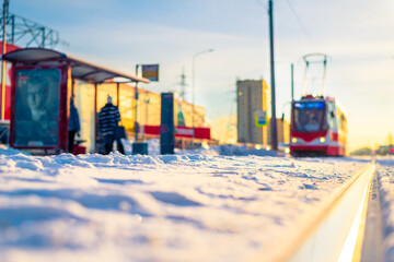 Tram stop with people. Sunrise on a winter morning. The tram is moving forward. Snow-covered road. Focus on rail. Close up view from ground level.