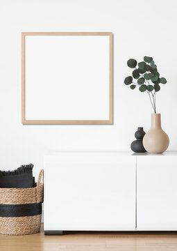 Empty square frame mockup in modern minimalist interior with plant in trendy vase and woven basket on white wall background. Template for artwork, painting or poster