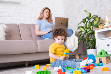 Mother freelancer use laptop sits on couch, kid daughter plays with toys on floor. Working from home