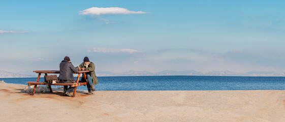 Old men sits on a bench by the sea. One man suggesting a book the other man. They are retired people. Living peaceful, quiet seaside town.	Empty space for text. Cop paste, web banner.
