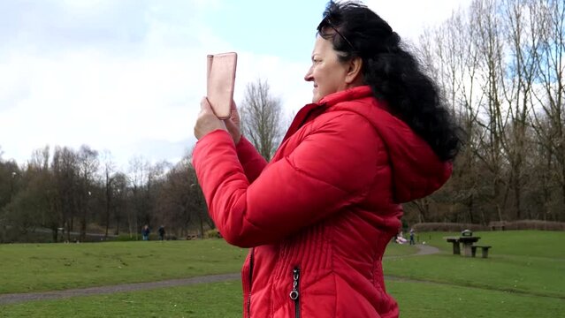Handheld camera following mature long hair woman in red jacket walking in Moses Gate country park in spring and taking pictures with her smartphone, England, United Kingdom.