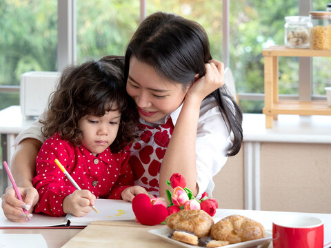 Pretty young Asian mom and adorable mixed race daughter sitting in the kitchen, drawing and making card for Valentine's Day, Mother's Day.