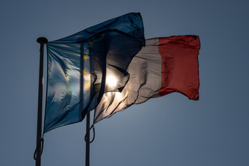 drapeaux de la France et de l'Europe côte à côte qui bougent dans le vent. On voit le soleil derrière les drapeaux par transparence. Ciel bleu en arrière plan