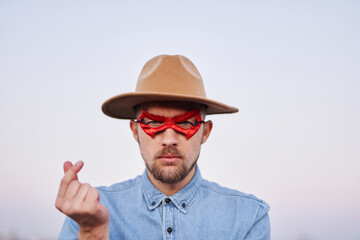 Cute bearded caucasian man in red superhero mask and jeans shirt posing with serious face in front of the camera. Male showing korean heart or mini heart gesture outdoor at sunset with urban view