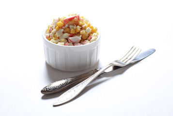 Salad with crab, egg and corn in a white bowl next to a fork and knife on a white background 