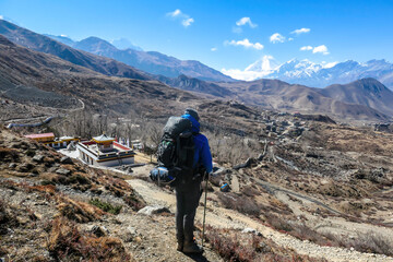 A man hiking through Himalayan valley, located in Mustang region, Annapurna Circuit Trek in Nepal. He is having a short break, supporting on hiking sticks and enjoying the view on small village