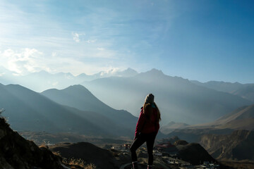 A girl wearing pink jacket standing on a top of a Himalayan mountain and enjoying the misty range spreading in front of her. Muktinath, Nepal. The sunlight is nicely marked. Freedom and achievement.