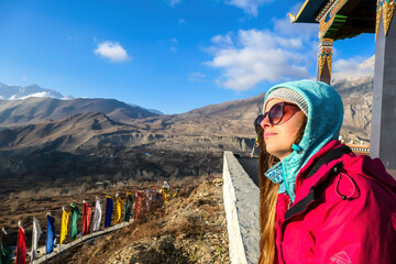 A girl wearing pink jacket sitting on a wall on a top of a mountain and enjoying the Himalayan range spreading in front of her. The woman enjoys sunbeams on her face. Freedom and achievement.