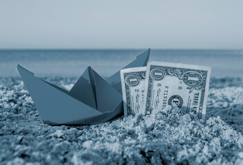 Orange paper boat and two paper dollar bills half buried in sand on sandy beach near sea. Small...