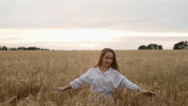 A young girl happily walking in slow motion through a field touching with hand wheat ears. Beautiful carefree woman enjoying nature and sunlight in wheat field at incredible colorful sunset