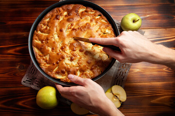 An appetizing apple pie in a baking dish stands on a wooden table. Human hands cut a piece of pie. 