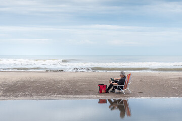 Mature adult male sitting on a beach chair pouring water in a mate all reflected in the water and...