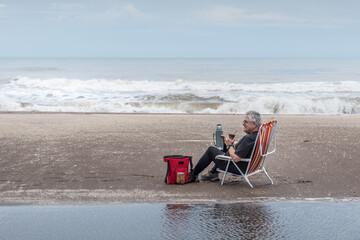 Mature man with gray hair and glasses sitting on a beach chair drinking mate all reflected in the...