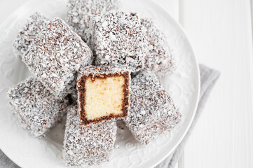 Traditional Australian Lamington cake in chocolate glaze and coconut flakes on a white plate on a white wooden background with a cup of tea. Selective focus.