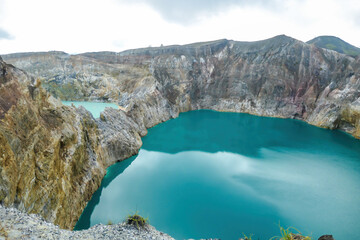 View on the Kelimutu volcanic crater lakes in Flores, Indonesia. Lakes are shining with many shades of turquoise and blue. Sun shines through clouds. Barren and sharp slopes of the volcanic crater