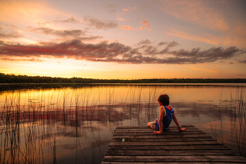 Naklejka na ściany i meble A young boy sits at the end of a dock and watches a summer sunset