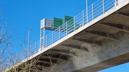 A gantry over a concrete road bridge showing road signs shot from below against a cloudless blue sky.