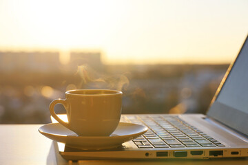 Coffee or tea cup with steam smoke on laptop against the window with sunshine. Cozy workplace in home office, concept of remote work