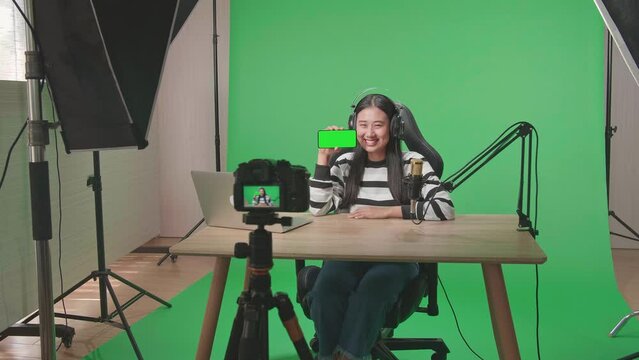 A Camera Recording A Smiling Asian Woman With Headphone And Computer Showing Green Screen Mobile Phone In Front Of Green Screen Background With Professional Light Equipment
