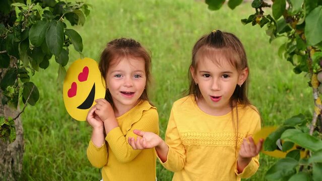 Girls in the garden under a tree hide behind cardboard faces of happy emoticons. world smile day