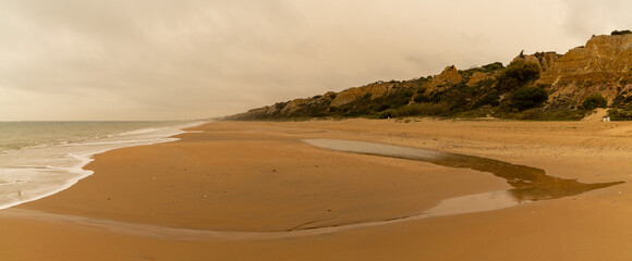 panorama view of a beautiful long and empty beach with shore break and high sand dunes behind and sahara dust in the air