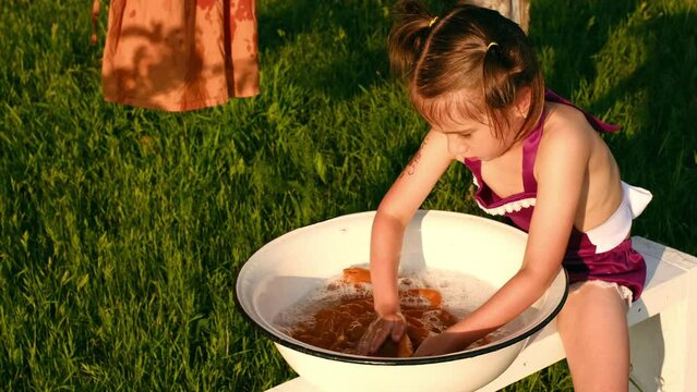 The girl is sitting on a bench in the garden and trying to laundry her shirt in a basin with powdered soap. Stain remover for colored clothes. manual washing