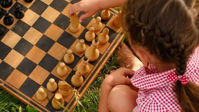A child makes the first move in chess with a knight on an old scratched aged wooden chessboard. Girl playing chess in the garden on the grass, close-up